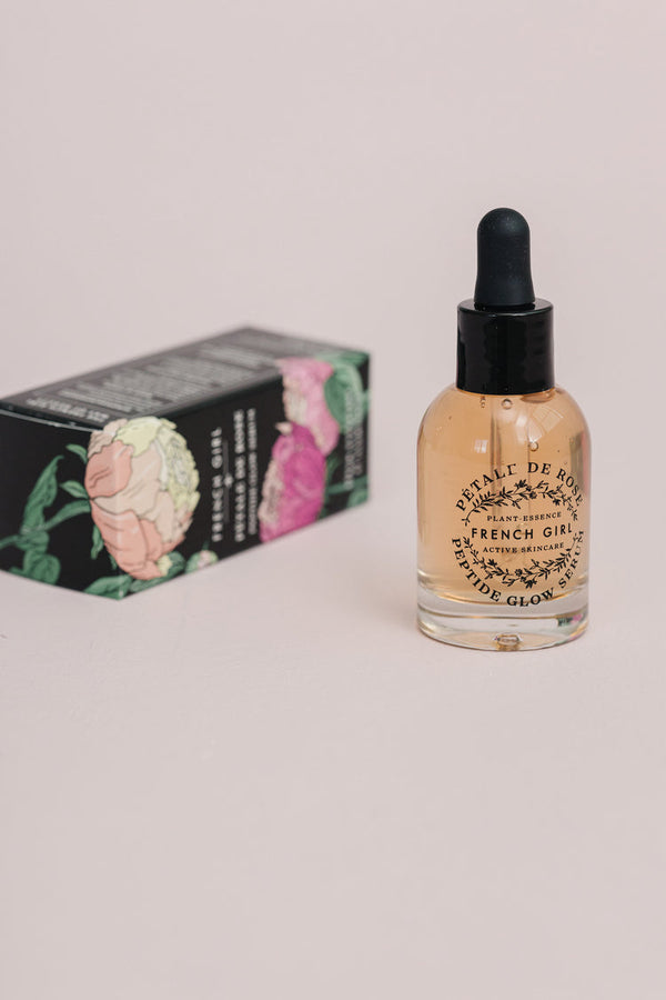 Clear glass dropper bottle of Peptide Glow Serum | French Girl with light amber liquid, standing beside a black box decorated with colorful floral illustrations for skincare use.