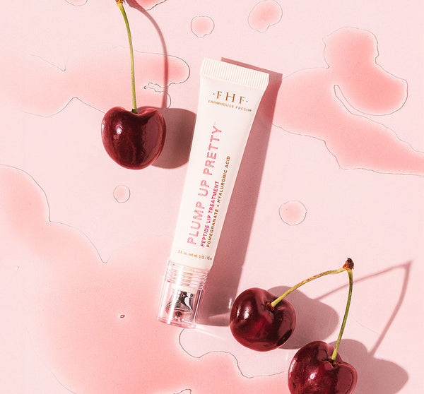 Plump Up Pretty Peptide Lip Treatment | Farmhouse Fresh tube on a pink surface surrounded by fresh dark cherries, highlighting skincare and lip care wellness.