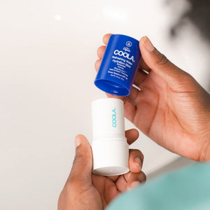 Person holding a white and blue tube of Refreshing Water Stick SPF 50 | COOLA skincare product with the cap removed, showcasing the hydration stick for body care.