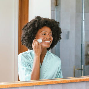 Woman in a light blue robe applying Refreshing Water Stick SPF 50 | COOLA to her face in a bright bathroom mirror, demonstrating skincare and sun protection.