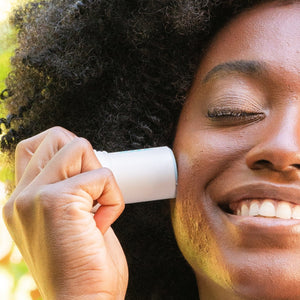 A smiling person with curly hair applies Refreshing Water Stick SPF 50 | COOLA to their cheek, showcasing easy-to-use skincare for sun protection and wellness.