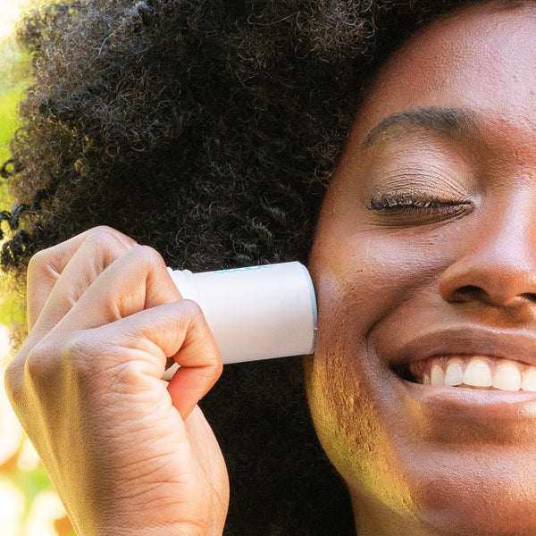 A smiling person with curly hair applies Refreshing Water Stick SPF 50 | COOLA to their cheek, showcasing easy-to-use skincare for sun protection and wellness.
