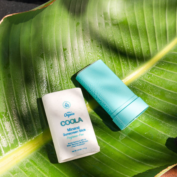 Mineral Stick SPF 50 | COOLA sunscreen stick in white and blue packaging displayed on a large green tropical leaf with natural sunlight and water droplets, emphasizing skincare and sun protection.