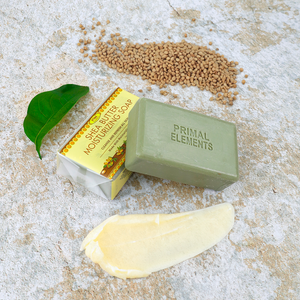 Luxury Shea Butter Moisturizing Soap | Primal Elements bar with natural ingredients beside a green leaf, moisturizing cream, and seeds on a textured surface representing skincare and body care.