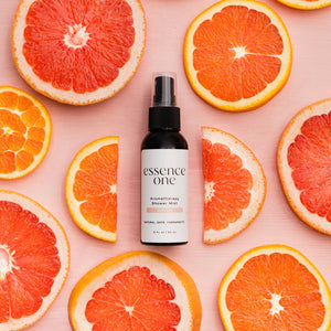 A bottle of Aromatherapy Shower Mist | Essence One surrounded by fresh sliced citrus fruits including grapefruit and orange on a pink background, highlighting a refreshing wellness skincare product.