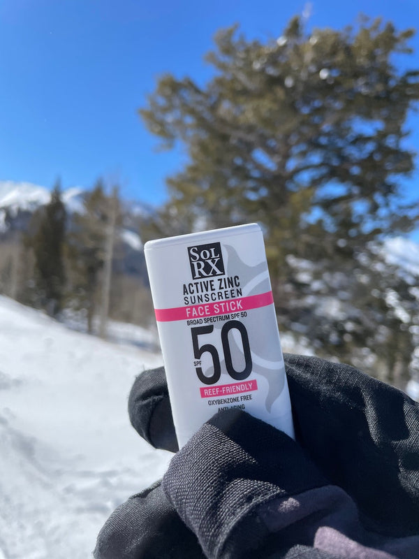 Person wearing black gloves holding Zinc Sunscreen Stick for Face | SolRX Sunscreen with SPF 50 outdoors in a snowy landscape with trees and a clear blue sky in the background.