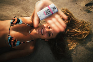 A woman lying on sand wearing a colorful bikini, holding and showing a Zinc Sunscreen Stick for Face | SolRX Sunscreen with SPF 50, smiling and enjoying skincare in the sun.