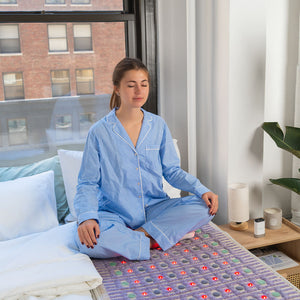 Woman in blue striped pajamas meditating while sitting cross-legged on the TAJ-Mat™ Large 8030 Firm - Photon PEMF (Right/Standard) Inframat Pro® | HealthyLine wellness device in a bright bedroom.