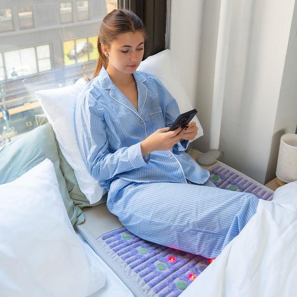 A woman in blue striped pajamas relaxing on a bed using her phone while lying on the TAJ-Mat™ Large 8030 Firm - Photon PEMF (Right/Standard) Inframat Pro® | HealthyLine wellness device for body care and recovery.