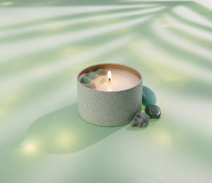 Bloom Candle Tin 6 oz | Aluminate Life burning with decorative stones on top, placed on a soft green surface with natural light and leaf shadows for a calming wellness ambiance.
