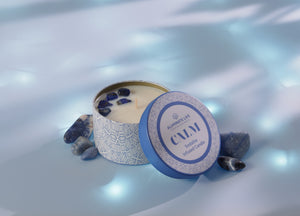 Calm Candle Tin 6 oz | Aluminate Life in a decorative blue and white tin surrounded by blue stones, creating a serene and relaxing atmosphere.