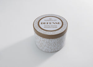 White and gold patterned tin candle container labeled Defense Candle Tin 6 oz | Aluminate Life, featuring a smoky quartz infused candle inside, placed on a white surface.