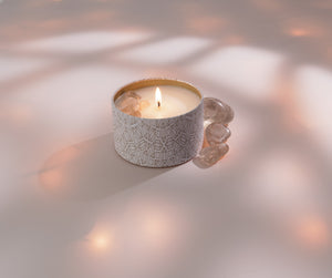 Lit candle in a decorative patterned tin with translucent stones beside it, showcasing the Defense Candle Tin 6 oz | Aluminate Life in a calming wellness setting.