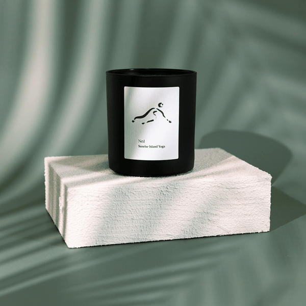 Black candle with minimalistic white label depicting a yoga pose, placed on a textured white block against a soft green background, featuring No. 1 Sunrise Island Yoga (Bali) Candle | Lucky Owl.