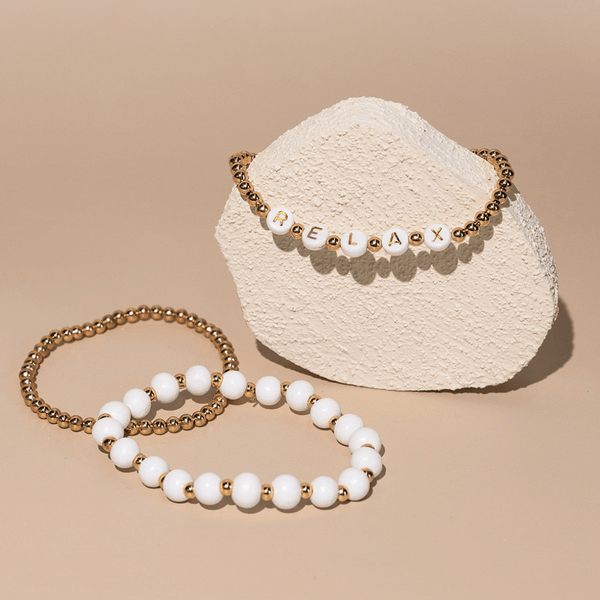 Wooden Beaded + Gold Bead 3 Bracelet Set - Relax | Lucky Owl displayed on a textured beige stand and flat surface, featuring white and gold round beads with letter beads spelling "RELAX" on one bracelet.