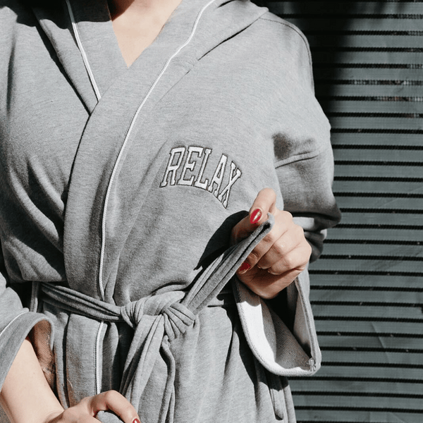 Close-up of a person wearing a cozy grey robe tied at the waist with "RELAX" embroidered on the chest, showcasing the Limited Edition RELAX Collegiate Robes | Lucky Owl for ultimate comfort and wellness.