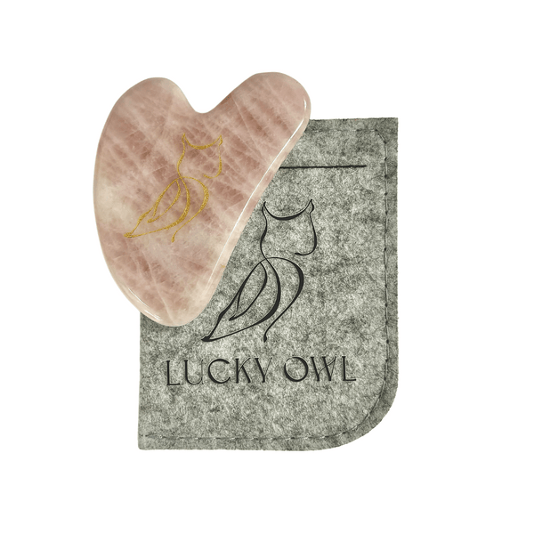 Gua Sha Kit | Lucky Owl with a heart-shaped rose quartz stone engraved with a gold owl logo, accompanied by a gray felt pouch featuring the Lucky Owl brand name and owl outline.