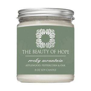 Rocky Mountain 8oz Candle | The Beauty of Hope in a clear glass jar with a silver lid, featuring a green label with white text and a floral emblem, scented with applewood, peppercorn, and oak.