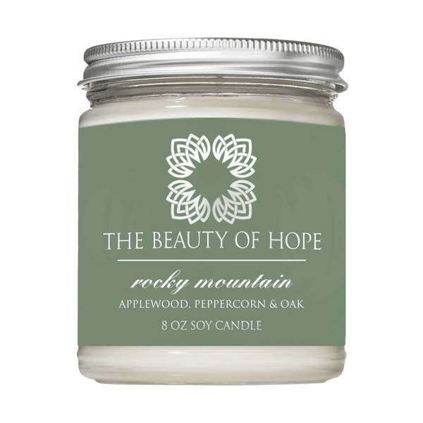 Rocky Mountain 8oz Candle | The Beauty of Hope in a clear glass jar with a silver lid, featuring a green label with white text and a floral emblem, scented with applewood, peppercorn, and oak.