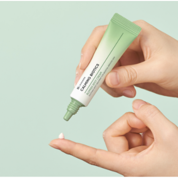 A close-up of hands holding and applying Calming Biotics Blemish Spot Cream | Leaders, with a small amount of cream dispensed on a fingertip against a soft green background.
