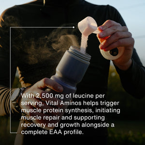 Person in outdoor setting pouring a scoop of Vital Aminos powder into a shaker bottle, highlighting the supplement’s muscle repair and recovery benefits with 2,500 mg leucine per serving.