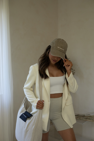 Woman wearing a white blazer, white crop top, and light gray shorts posing indoors while holding a large natural linen tote bag, promoting the Linen Acupressure Mat | Vie Healing.
