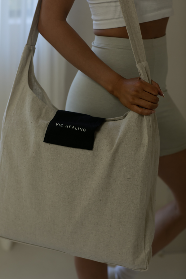 A person holding a large natural linen tote bag with a black label reading Vie Healing, styled casually in light-colored activewear, representing the Linen Acupressure Mat | Vie Healing.