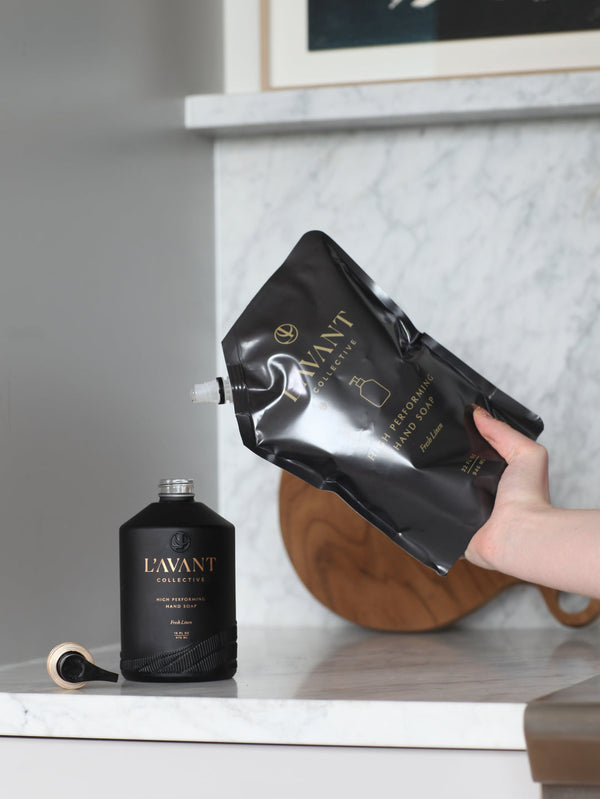 Refilling a sleek black bottle of High Performing Hand Soap - Fresh Linen | L'AVANT Collective on a marble countertop in a modern kitchen setting, highlighting premium skincare and body care.