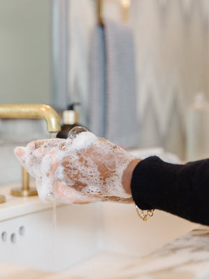 Hands covered in foamy soap lather being washed under running water at a modern bathroom sink, demonstrating use of High Performing Hand Soap - Fresh Linen | L'AVANT Collective for skincare and body care.