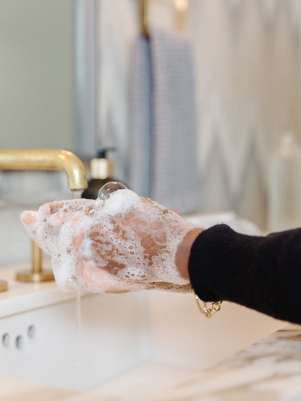 Hands covered in foamy soap lather being washed under running water at a modern bathroom sink, demonstrating use of High Performing Hand Soap - Fresh Linen | L'AVANT Collective for skincare and body care.