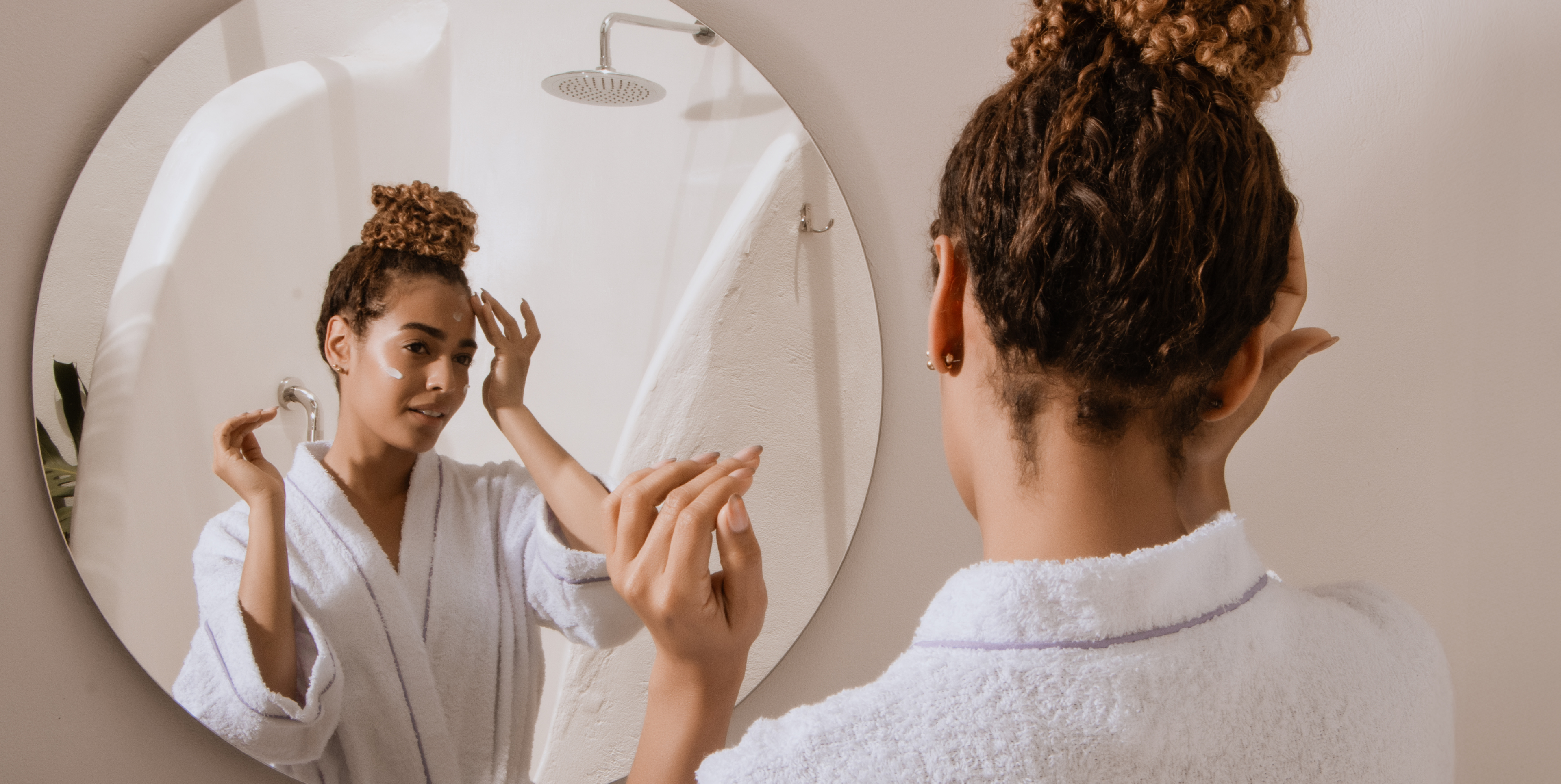 A woman with curly hair in a white bathrobe stands in front of a round mirror, gently touching her face in a softly lit bathroom setting.