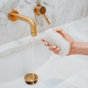 A hand holding the Konjac Walnut Exfoliating Body Sponge | Bathorium under running water from a gold faucet over a white sink with marble backsplash.