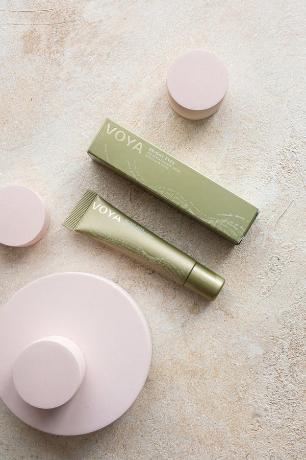 Green tube and matching box of Bright Eyes Regenerating Eye Cream | VOYA displayed on a textured beige surface with soft pink circular props, representing skincare and wellness.