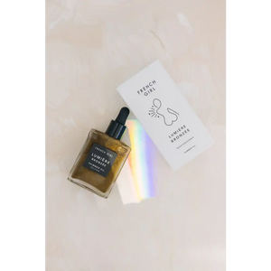 Bronzée Shimmer Oil| French Girl bottle with dropper and its white packaging featuring minimalist line art, set on a light surface with a subtle rainbow reflection.