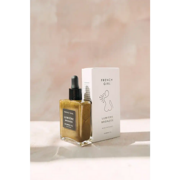 Bronzée Shimmer Oil| French Girl in a clear glass dropper bottle with golden shimmer next to a white box featuring minimalist line art, set against a soft neutral background.