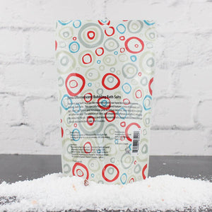 Back view of Detox Bubbling Bath Salts | Primal Elements in a colorful circular patterned pouch, resting on a pile of white bath salts against a light brick background.