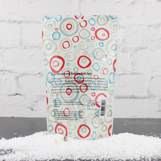 Back view of Detox Bubbling Bath Salts | Primal Elements in a colorful circular patterned pouch, resting on a pile of white bath salts against a light brick background.