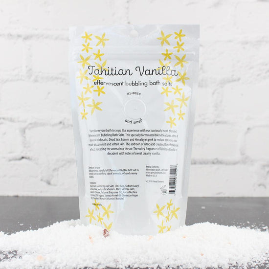 Tahitian Vanilla Bubbling Bath Salts | Primal Elements package surrounded by white bath salts on a dark surface against a white brick background, showcasing skincare and wellness body care.