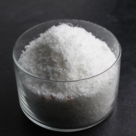Clear glass container filled with white crystalline Tahitian Vanilla Bubbling Bath Salts | Primal Elements, displayed on a dark surface for skincare and wellness use.