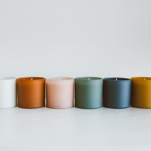 A row of six glass jar candles in varying muted colors including white, terracotta, blush pink, sage green, slate blue, and mustard, featuring the Evergreen + Eucalyptus Dignity Series Glass Jar Soy Candle | Calyan Wax Co. - 5.3 oz.