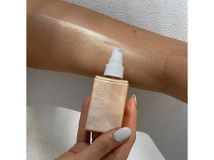 Hand holding a gold bottle of Body Shimmer | NUDA next to a forearm with glowing, radiant skin showcasing a smooth, luminous shimmer effect from the body care product.