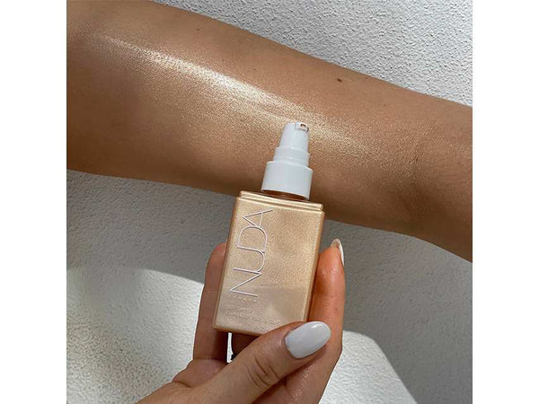 Hand holding a gold bottle of Body Shimmer | NUDA next to a forearm with glowing, radiant skin showcasing a smooth, luminous shimmer effect from the body care product.