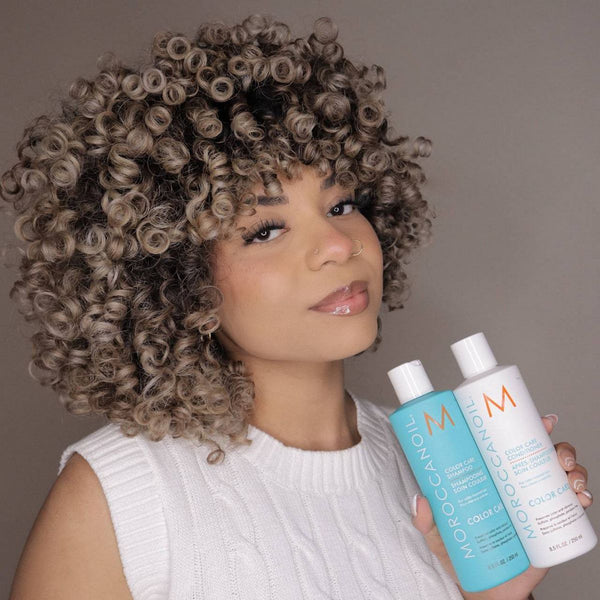Woman with curly hair holding two bottles of Color Care Shampoo - Travel | Moroccanoil, promoting hair care and wellness.