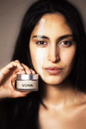 Woman with glowing skin holds a jar of Get Glowing Illuminating Clay Mask | VOYA while a streak of the clay mask is applied vertically on her face, showcasing skincare benefits.