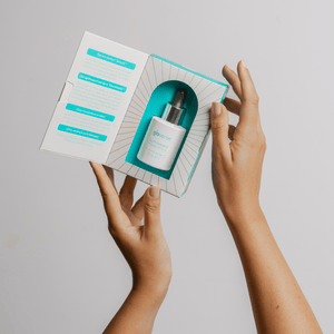 Hands holding an open box containing a dropper bottle of LUMINISCENT Radiance Facial Oil | Glo910, displayed against a light gray background.