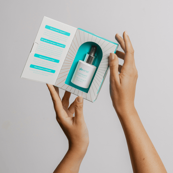 Hands holding an open box containing a dropper bottle of LUMINISCENT Radiance Facial Oil | Glo910, displayed against a light gray background.