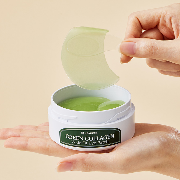 A hand holding a transparent spatula lifting a green collagen eye patch from an open white container labeled Green Collagen Wide Fit Eye Patch | Leaders, with another hand supporting the container.