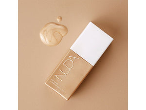 Sleek gold bottle of Body Shimmer | NUDA with a shimmer liquid drop on a beige background, highlighting a radiant, glowing body care product for skincare and wellness.