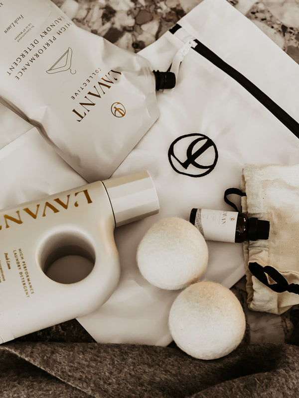 Laundry Detergent Refill Pouch | L'AVANT Collective featuring a modern beige refill pouch, a matching bottle, two wool dryer balls, and a small essential oil bottle on a white fabric background.