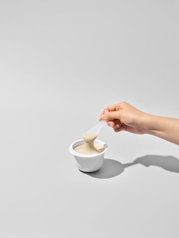 Hand holding a spatula scooping creamy skincare product from a white cup, showcasing 3X Boosting Modeling Mask - Vitamin | Leaders for a smooth skincare routine.